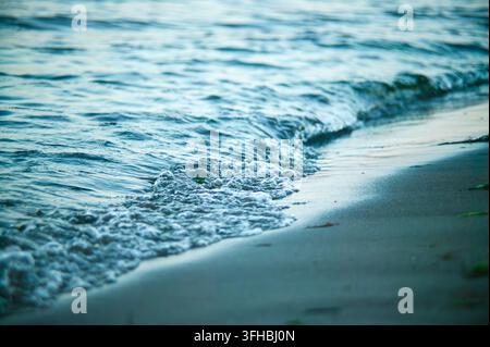 L'acqua dell'oceano si infrange in piccole onde con schiuma e bolle, lavando delicatamente sulla sabbia bagnata e scura della spiaggia, creando una natura tranquilla e serena Foto Stock