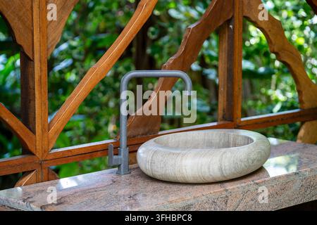 Lavandino moderno in pietra e rubinetto in interno esterno in legno. Elegante lavabo rotondo in pietra e rubinetto grigio opaco su piano di lavoro in granito davanti al legno Foto Stock