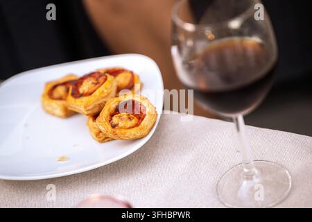 Spuntini salati di pasta sfoglia sono serviti su un piatto bianco accanto a un bicchiere di vino rosso, creando un'atmosfera invitante per un incontro sociale informale o una festa Foto Stock