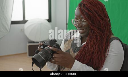 Donna tiene la fotocamera e controlla le impostazioni in studio con schermo verde, softbox e finestra visibili; concentrazione creatività. Foto Stock