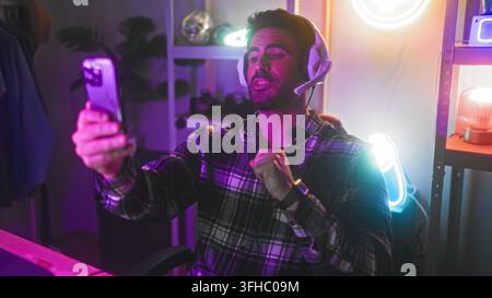 Giovane uomo con auricolari tiene uno smartphone e porta le dita sulle labbra per indicare silenzio in uno studio con luci al neon e scrivania da gioco; coinvolgimento giocoso. Foto Stock
