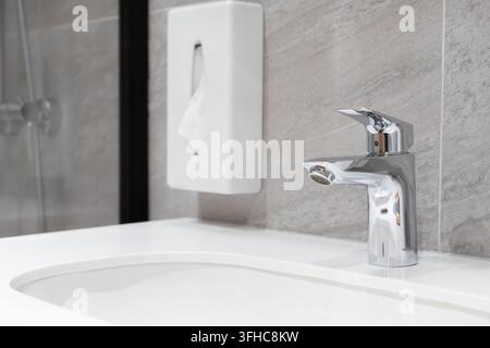 Il rubinetto miscelatore in acciaio inossidabile è posizionato su un lavabo contemporaneo all'interno di un bagno minimalista, dove sottili texture di pietra e attrezzature ordinate rappresentano Foto Stock