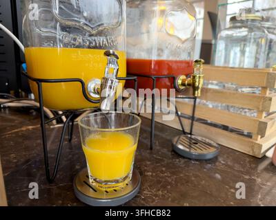Vista dall'alto di un distributore di succo in vetro che versa un succo d'arancia vivace in un bicchiere, con un distributore di succo rosso nelle vicinanze, ideale per la colazione in hotel o il catering Foto Stock