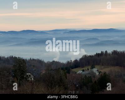 Vasta veduta aerea che cattura strati di montagne azzurri e sfumati avvolti nella nebbia, con fumo che si alza da un villaggio rurale sottostante, riflettendo la bellezza naturale Foto Stock