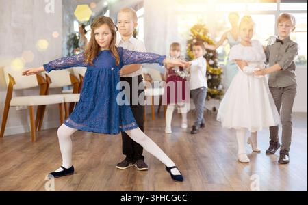 Preadolescenti entusiasti, ragazze e ragazzi in abiti eleganti che praticano passi di danza di coppia in uno studio adornato di decorazioni natalizie, sotto Gu Foto Stock