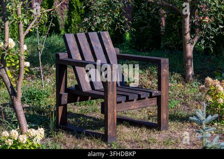 Poltrona in legno fatta a mano in giardino verde, primo piano. Poltrona in legno marrone scuro in stile Adirondack in piedi sull'erba in un giardino soleggiato circondato da Tr Foto Stock