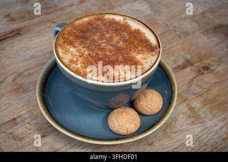 Chai latte cosparso di cannella in una tazza blu salvia con piattino e due piccoli biscotti amaretti su un tavolo di legno Foto Stock