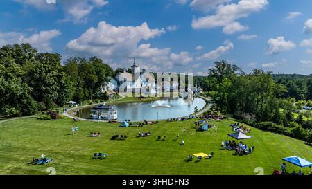 Molte persone si radunano sull'erba di un parco in una giornata di sole. Uno stagno è visibile al centro, con edifici sullo sfondo. Tende e sedie sono sistemate intorno all'area per il relax. Foto Stock