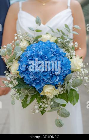 Sposa con bouquet, in un ambiente all'aperto, forse luogo di matrimonio, con abito da sposa bianco, ortensie blu, rose bianche, gipsofila, da un primo piano Foto Stock