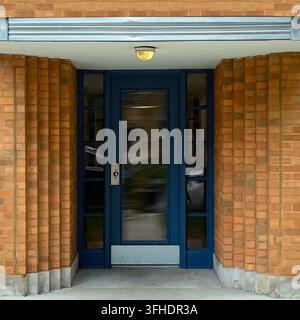 Porte blu in stile Streamline Moderne con vetri laterali stretti sono incorniciate da pilastri in mattoni arancioni sporgenti sotto una tettoia chiara e una luce a soffitto. Foto Stock