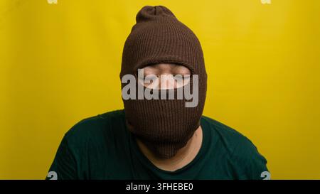 Giovane uomo asiatico con passamontagna marrone in piedi con gli occhi chiusi su uno sfondo giallo vivace. L'individuo mascherato proietta un'aura di pace Foto Stock