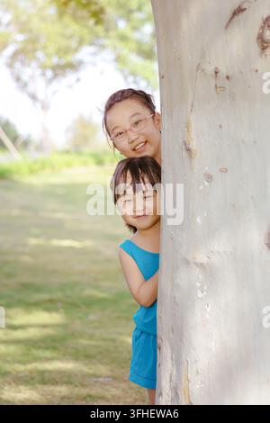 Due adorabili sorelle asiatiche che giocano a nascondino dietro un grosso tronco d'albero nel parco, sorridendo e sbirciando la fotocamera, infanzia gioiosa all'aperto estate a Foto Stock