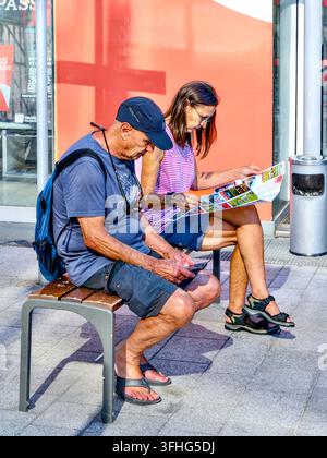 Coppia di uomo e donna, vestiti in modo casual, seduti su una panchina all'aperto cercando informazioni su mappa e smartphone – Tours, Indre-et-Loire (37), Francia. Foto Stock