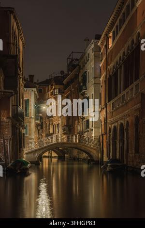 Vista di un canale tranquillo di notte, con un ponte in pietra ad arco, edifici storici con finestre che emanano una luce calda e piccole barche ormeggiate sull'acqua calma Foto Stock