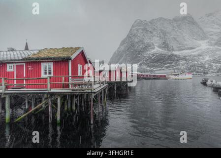 Vista delle tradizionali cabine rosse in legno rorbuer su palafitte sopra l'acqua con montagne innevate sullo sfondo a Lofoten, Nordland, Norvegia. Foto Stock