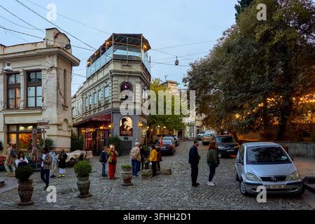 Le persone si radunano su una strada acciottolata nella piazza storica europea con vecchi edifici, ristoranti e auto durante il crepuscolo serale Foto Stock
