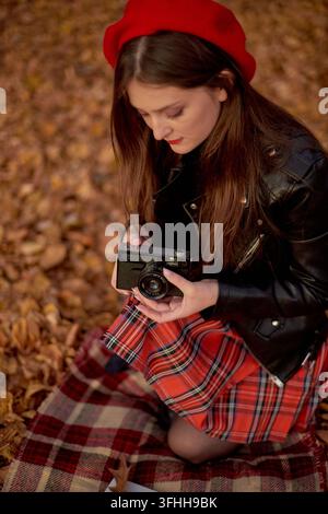Giovane donna con berretto rosso che ispeziona una fotocamera retrò durante un picnic autunnale Foto Stock