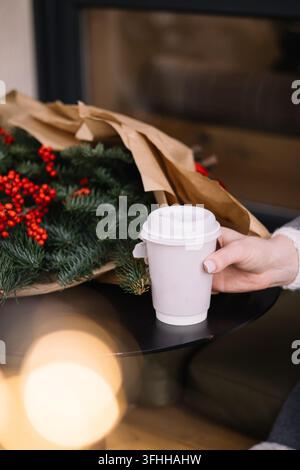 Mano femminile con una manicure chiara e curata che tiene una tazza di caffè da asporto in carta bianca con coperchio su un tavolo rotondo nero da caffè, accanto a un mazzo di fiori freschi Foto Stock