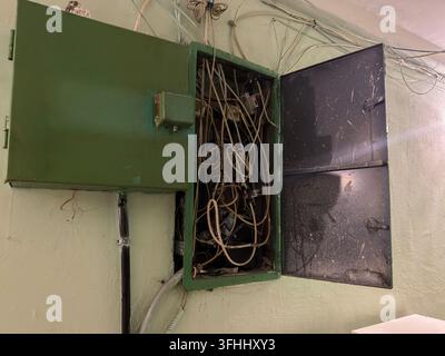 Disordine caotico di cavi elettrici esposti all'interno di un armadietto verde, che dimostra una gestione del cablaggio obsoleta e pericolosa e l'urgenza per pr Foto Stock
