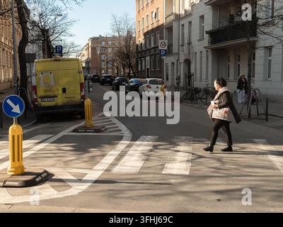 Poznań, Polonia – 6 marzo 2026: Pedone che attraversa una strada su un passaggio pedonale segnalato vicino a macchine parcheggiate e un furgone giallo, riflettendo la routine di dai Foto Stock