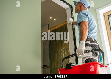 Un operaio che indossa guanti e occhiali di sicurezza controlla una cornice della porta mentre tiene una cassetta degli attrezzi accanto all'ingresso del bagno. Foto Stock