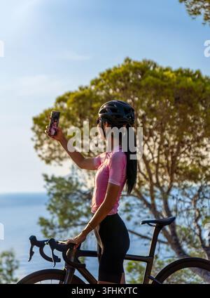 Una giovane ciclista scatta un selfie con il suo smartphone accanto alla sua bici da strada su una strada costiera vicino a Tossa de Mar, con il Mar Mediterraneo in Foto Stock