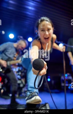 Giovane cantante con capelli scuri ed espressione intensa tende un microfono cablato verso la telecamera su un palco illuminato di blu accanto al chitarrista durante una performance dal vivo Foto Stock