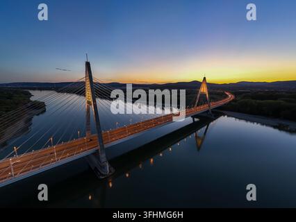 Vista aerea del ponte Megyeri sul Danubio a Budapest, Ungheria, illuminato nell'ora blu con traffico autostradale, riflessi sull'acqua e urba Foto Stock
