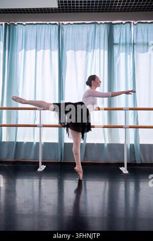 Giovane ballerina con capelli scuri allunga una gamba accanto alla sbarra di legno in uno studio di danza luminoso, in equilibrio sulla punta davanti alle tende blu con concentrazione Foto Stock