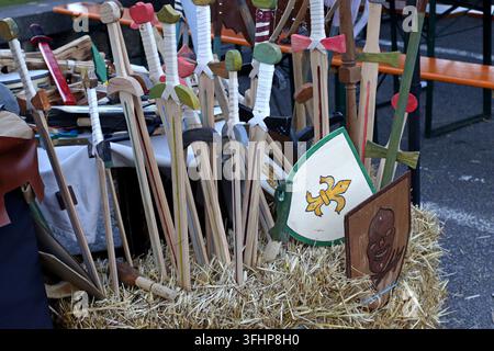 Collezione di spade e scudi giocattolo in legno infantili esposti nella paglia a una fiera medievale, manici colorati, dettagli intagliati e motivi araldici come t Foto Stock