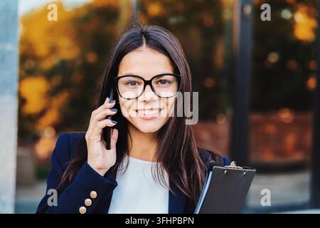 Donna d'affari ha una conversazione usando il telefono cellulare. Donna d'affari con occhiali e completo formale parla allegramente con un collega. Offi Foto Stock