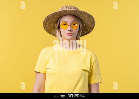 Ritratto di giovane bella donna bionda su sfondo studio giallo estate. Abbigliamento soleggiato e sicuro con occhiali da sole e cappello in paglia. Foto Stock