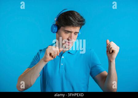 Giovane uomo affascinante con acconciatura alla moda che si diverte, balla, sorride con le cuffie in studio su sfondo blu. Musica, dan Foto Stock