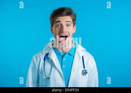 Medico stupito, scioccato, che dice WOW. Bel medico in camice bianco sorpreso verso la telecamera su sfondo blu. Foto di alta qualità Foto Stock