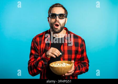 Giovane uomo con occhiali 3D che guarda un affascinante film horror e mangia popcorn su uno sfondo blu da studio. Foto Stock