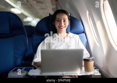 Giovane donna d'affari asiatica di successo siede in aereo a cabina e lavora da qualsiasi luogo. Volare in prima classe Foto Stock