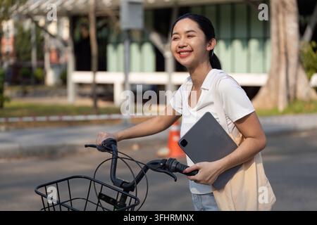 Una donna allegra tiene un tablet mentre cammina con la sua bicicletta, incarnando uno stile di vita che valorizza la sostenibilità e la tecnologia. Foto Stock