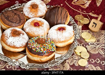 Ciambelle tradizionali per Hanukkah con gelatina, cioccolato e zuccherini su un vassoio d'argento accanto a un dreidel di legno e monete d'oro su una tovaglia rossa. Foto Stock