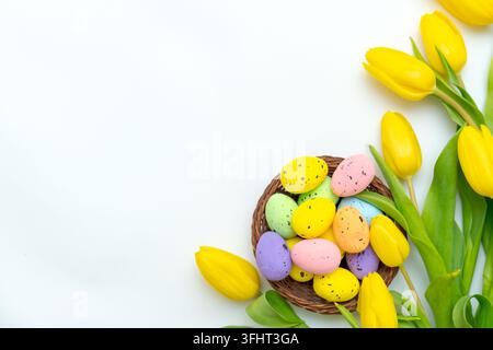 Uova di Pasqua colorate in un cesto di vimini con tulipani gialli su uno sfondo bianco. Foto Stock