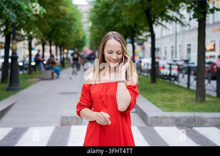 Donna d'affari attraente che parla su un telefono cellulare con un cliente in piedi su una strada della città durante il giorno in un vestito rosso. Donna caucasica che riesce Foto Stock