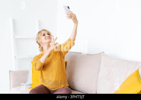 Donna caucasica sorridente seduta sul divano a casa, che scatta un autoportrato su moderno Foto Stock