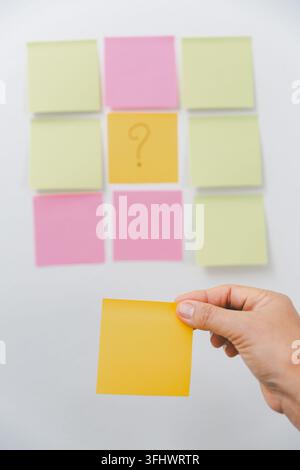 Mano che tiene un post-it giallo vuoto davanti a note colorate con un punto interrogativo, rappresentante il processo decisionale, l'incertezza, la risoluzione dei problemi, p Foto Stock