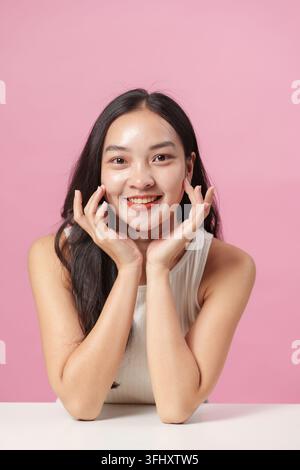 Una giovane donna con un sorriso luminoso posa con le mani sul viso, evidenziando la bellezza e la cura di sé su uno sfondo rosa. Foto Stock