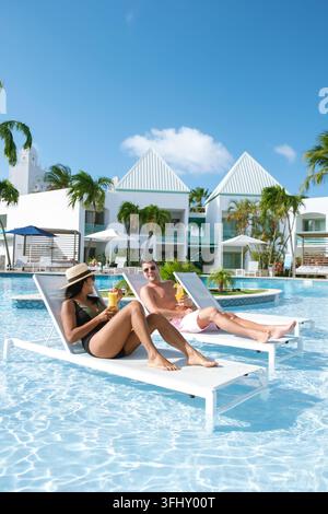 Due vacanzieri si rilassano su sedie a bordo piscina, gustando bevande rinfrescanti sotto il sole. Circondato da palme e da un'architettura vivace, questo tropicale Foto Stock