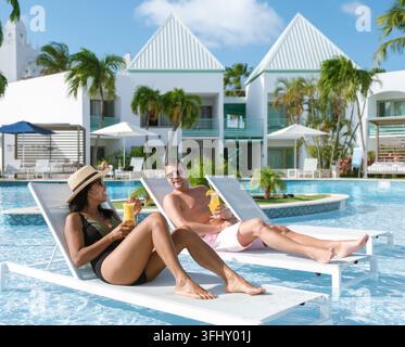 Due turisti si rilassano in una scintillante piscina ad Aruba, gustando cocktail tropicali sotto il sole. Il vivace paesaggio e le palme creano il buongiorno perfetto Foto Stock