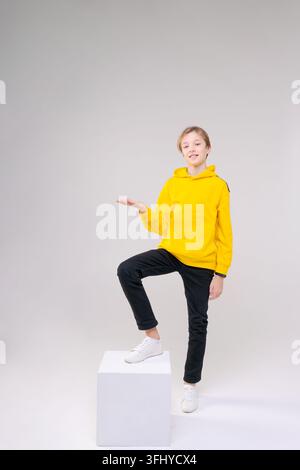 Giovane uomo bello, felice e sorridente in jeans neri, felpa giallo chiaro con cappuccio, si rallegra in posa su uno sfondo chiaro. Il conc Foto Stock