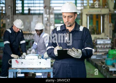 Capo squadra industriale in piedi in una fabbrica di produzione con operai che azionano macchinari sullo sfondo, rappresentando il lavoro di squadra e le pubbliche relazioni industriali Foto Stock