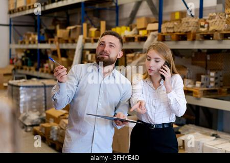 Responsabili della logistica professionali in un magazzino. Una donna parla con uno smartphone e punta all'inventario mentre l'uomo tiene gli appunti. Concetto di efficacia Foto Stock