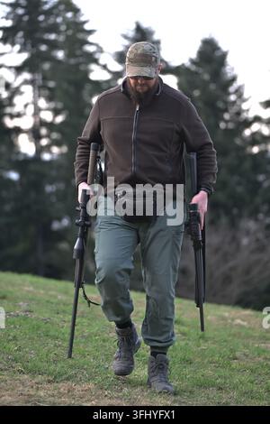 Cacciatore barbuto attraversa una radura erbosa della foresta portando due fucili e attrezzatura da esterno, indossa un cappello mimetico, una giacca tattica e stivali. Atmosferico Foto Stock
