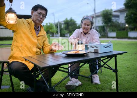 Coppia di anziani asiatici che si rilassano e gustano snack leggeri a un tavolo da campeggio nel loro giardino, condividendo un momento tranquillo all'aperto insieme Foto Stock